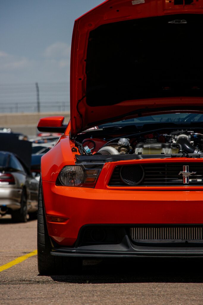 Ford Mustang arancione con cofano aperto in un evento automobilistico, vista frontale con motore elaborato e griglia iconica con logo del cavallo al galoppo.